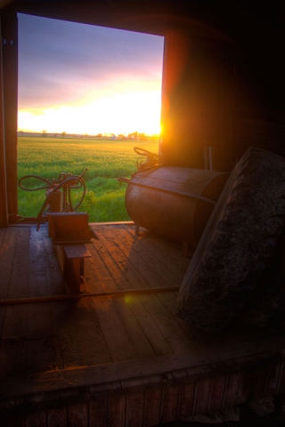 View from inside a dark room or building looking out through a window or doorway at a sunset over green fields.