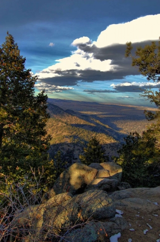 Elevated view of a valley or landscape from a rocky overlook, with dramatic clouds and blue sky above forested hills.