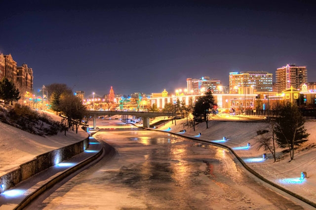 Night cityscape with a river or canal in the foreground, showing city lights reflected on the water's surface.