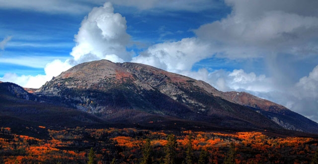 Mountain landscape with a large peak partially covered by clouds against a blue sky, with autumn foliage visible in the foreground.