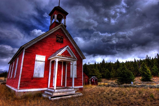 Small red schoolhouse or church with a bell tower under a dramatic cloudy sky in a rural setting.