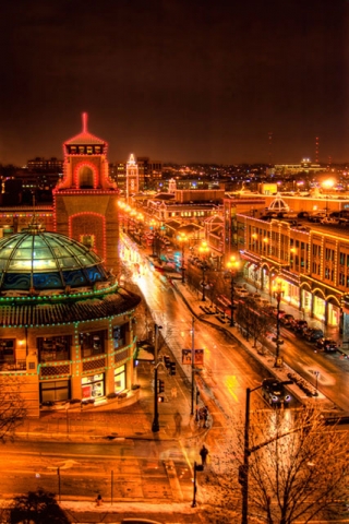 Nighttime cityscape showing illuminated historic buildings along a main street, possibly in a European or older American city, with a domed structure visible.