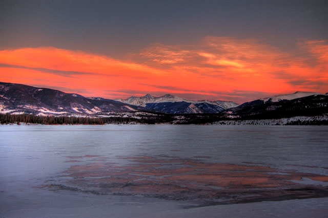 Winter sunset over a frozen lake or snow-covered landscape with mountains in the background and dramatic pink-orange sky.