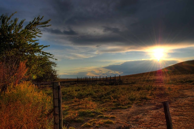 Sunset landscape with a fence line and autumn foliage under a dramatic cloudy sky.