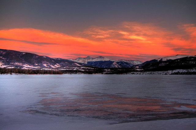 Sunset or sunrise over a snow-covered landscape or frozen lake with mountains in the background and a vivid orange-red sky.