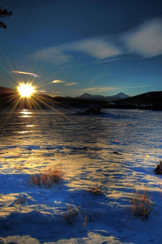 Sunset or sunrise over a frozen lake or snowy landscape with sunlight reflecting off the ice and mountains in the background.