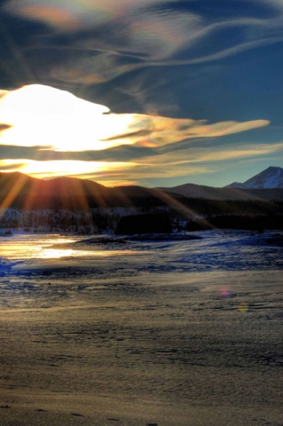 Sunrise or sunset over a winter landscape with light casting dramatic rays across snowy ground with mountains in the background.
