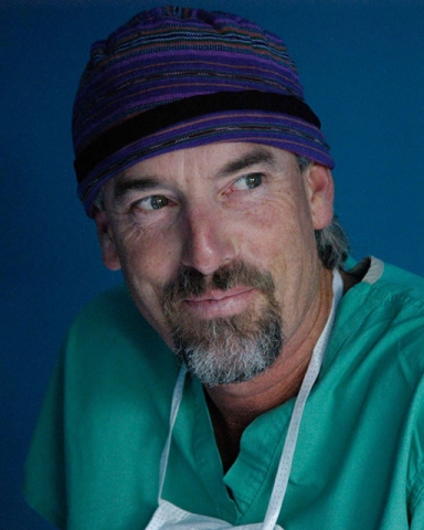 Portrait of a medical professional wearing a blue surgical cap and green scrubs, possibly a surgeon or doctor.
