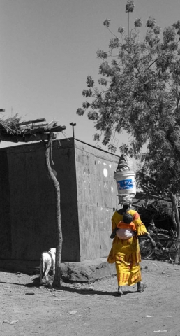 A person in yellow clothing carrying something on their head walking near a simple structure in a rural setting. The image is in black and white except for the person's clothing.