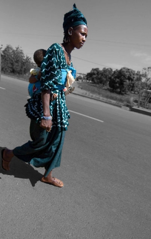 A person in colorful blue traditional dress walking barefoot on a road, carrying a child on their back. The background is in black and white while the person's clothing is in color.