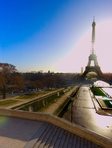 The Eiffel Tower in Paris viewed from a distance, with gardens or pathways in the foreground during sunset or sunrise.