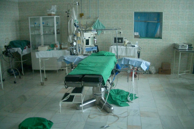 An operating room or medical facility with a surgical bed, medical equipment, and green surgical drapes.