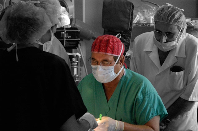 Medical personnel in surgical attire, with one wearing a red surgical cap, performing what appears to be a medical procedure.