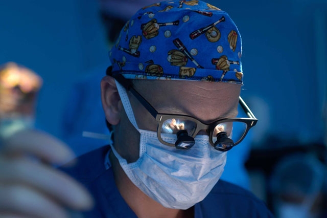 A medical professional wearing surgical glasses, a face mask, and a patterned surgical cap under blue medical lighting.