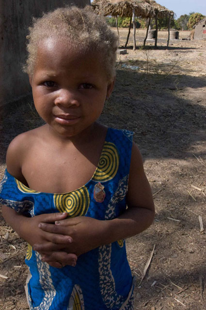 A young child standing outdoors in a patterned blue outfit. The child has lighter-colored hair and appears to be in an arid or rural setting.
