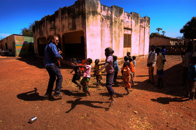 Several people, possibly children, playing or gathering outside what appears to be a building with reddish/pink walls under a blue sky.