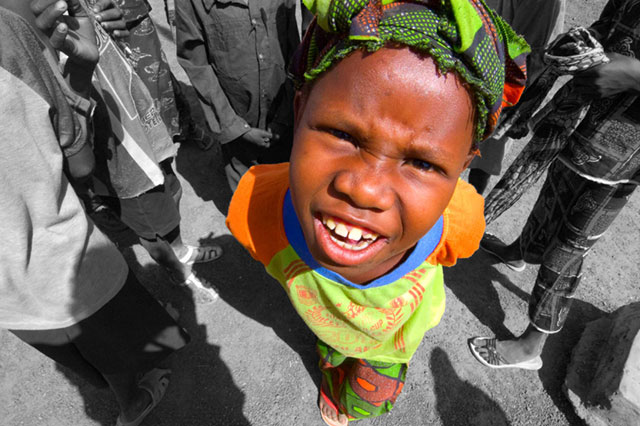 Image of a child with a bright yellow/orange garment or scarf, smiling at the camera. The background appears to be in black and white while the child's clothing is in color.