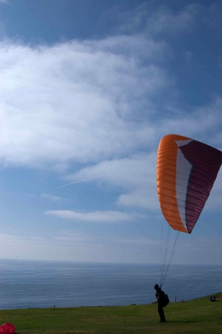 A person paragliding with an orange and dark-colored parachute against a blue sky over what appears to be a shoreline.
