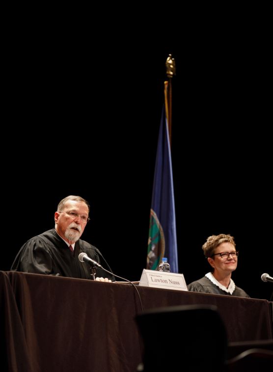 two judges sit at a long table with a black table cloth with microphones in front of them