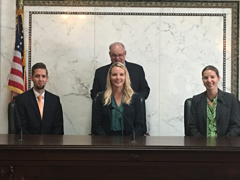 Ashley Akers sitting behind a podium with three other people in a courtroom.
