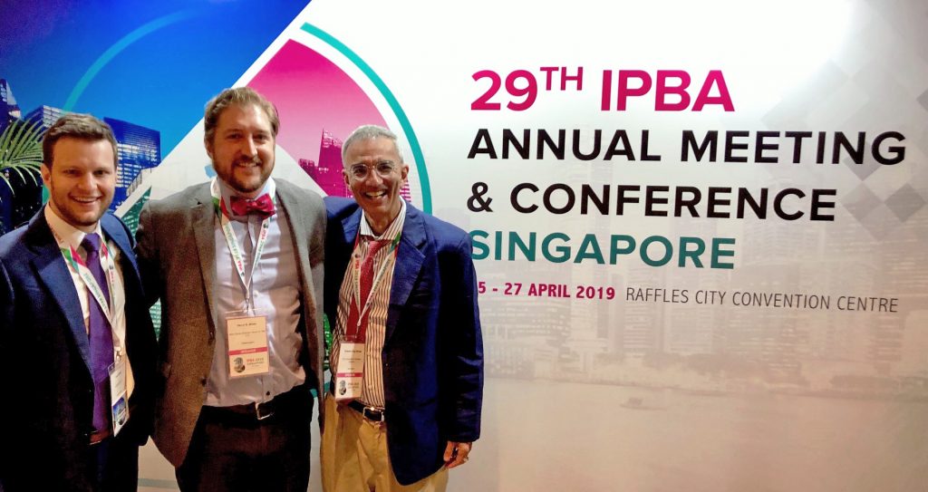 3 people standing next to the board of the Inter-Pacific Bar Association’s annual meeting in Singapore