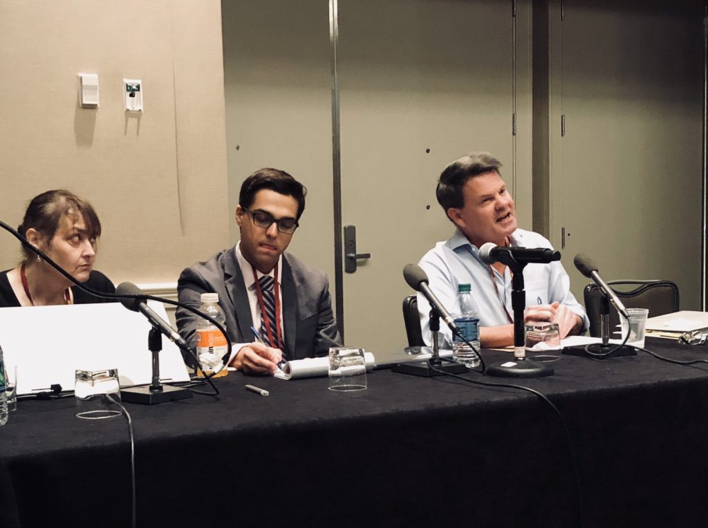 Three people behind a long table with papers and microphones on it