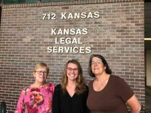 Three women smiling in front of a Kansas Legal Services sign