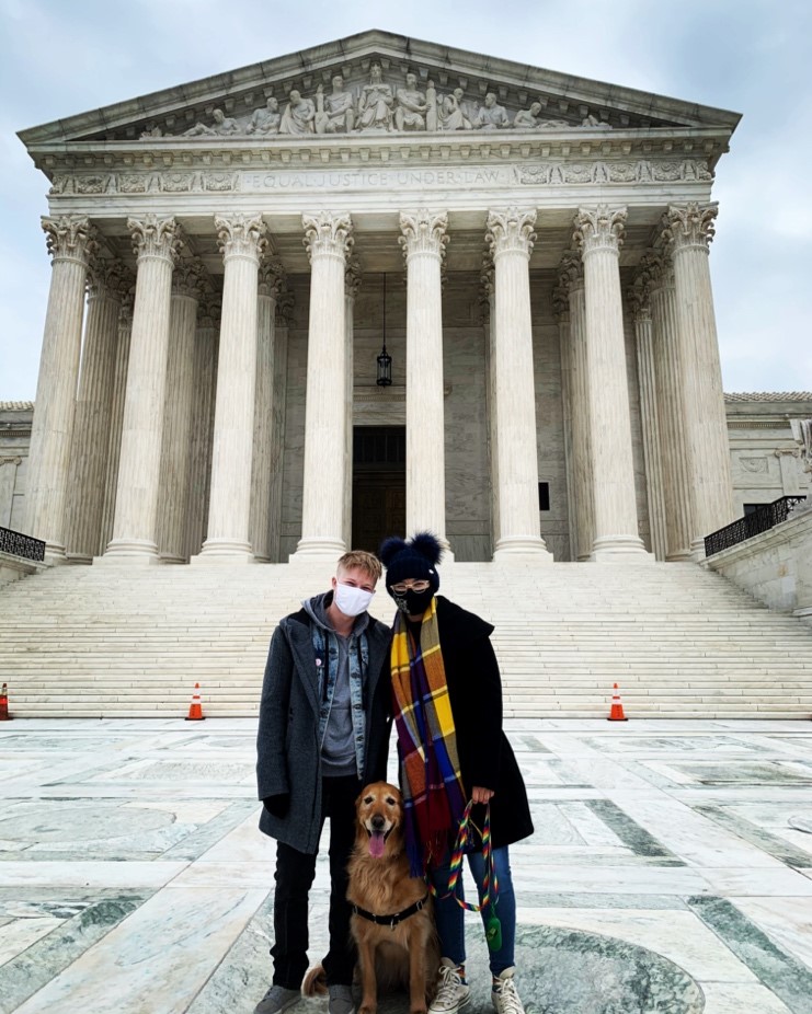 Two people standing together with a golden retriever in front of a white building with pillars.