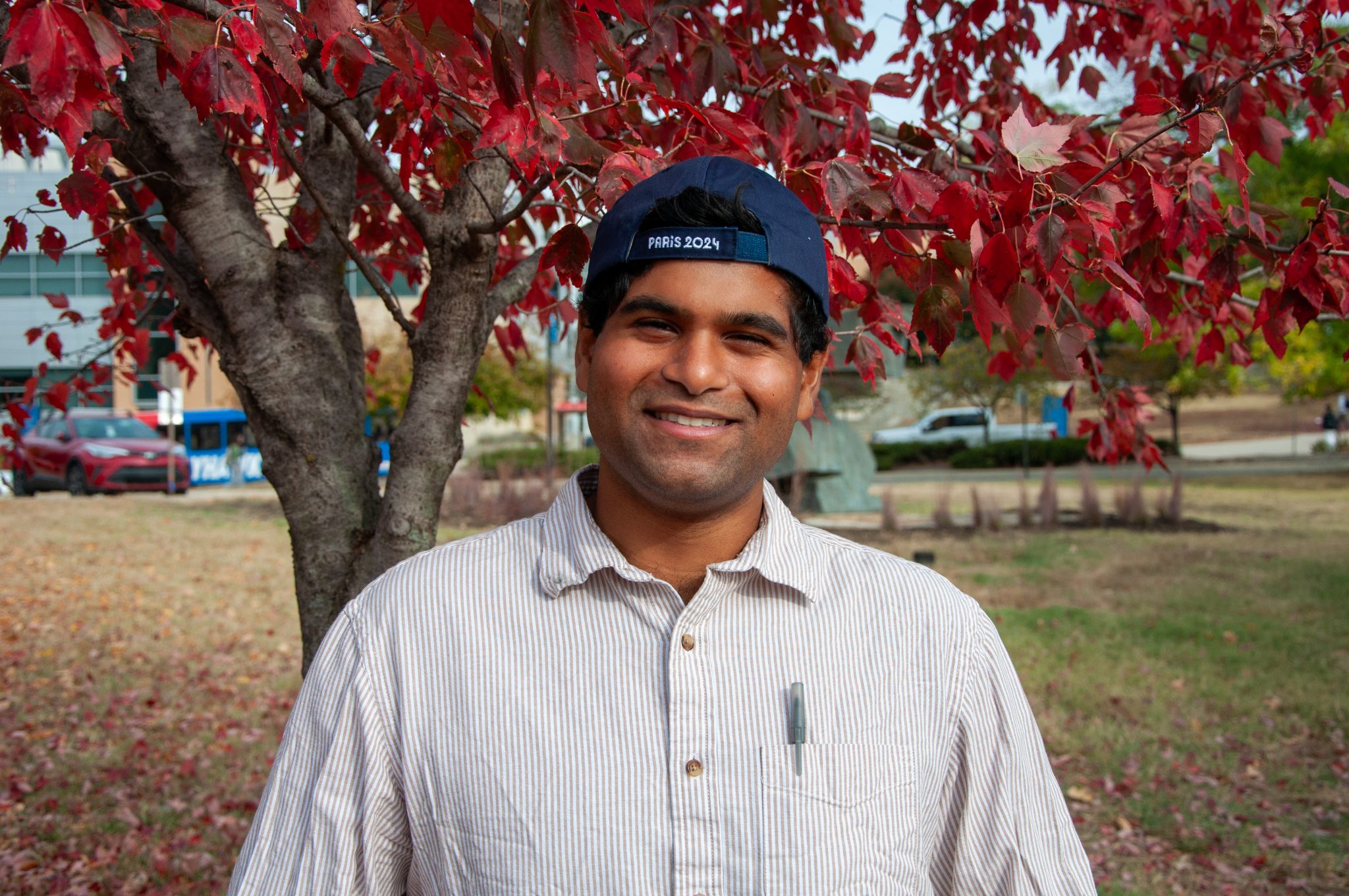 Sidharth Mohan wearing a backwards baseball cap, smiling in front of a tree with red leaves on campus