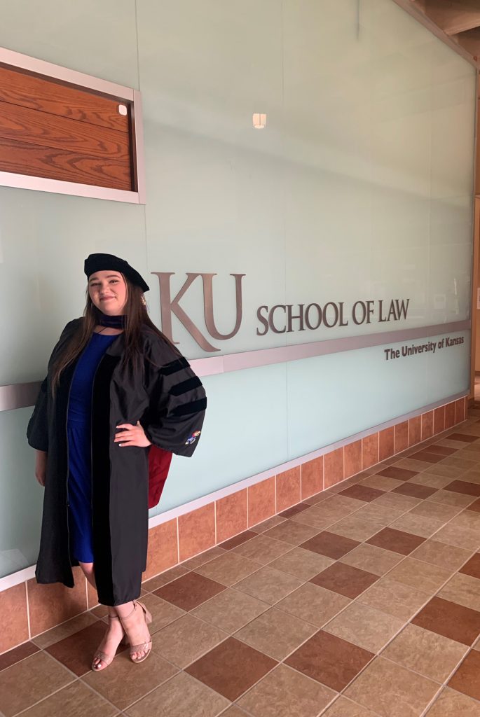 Ashlyn Shultz in graduation regalia in front of the KU School of Law wall sign