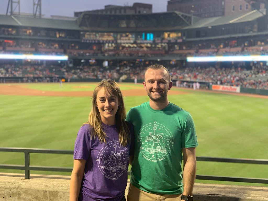 two people posing by a railing at the edge of a baseball diamond