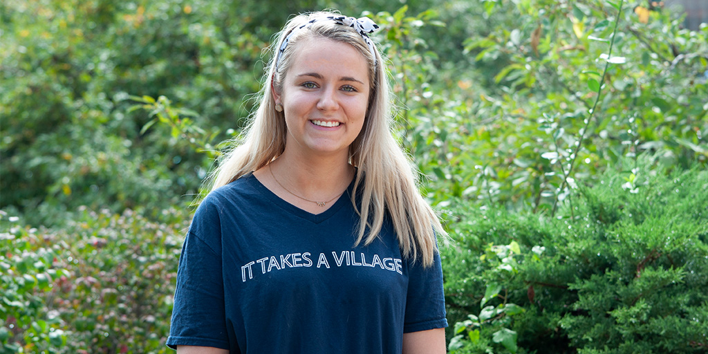 Ashley Golledge wearing a shirt that says "It Takes a Village" in front of vegetation