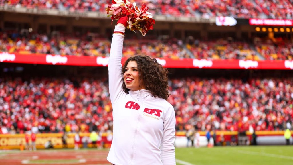 Noell Hinsley in Chiefs jacket holding pompom above head with crowded stadium seating in background