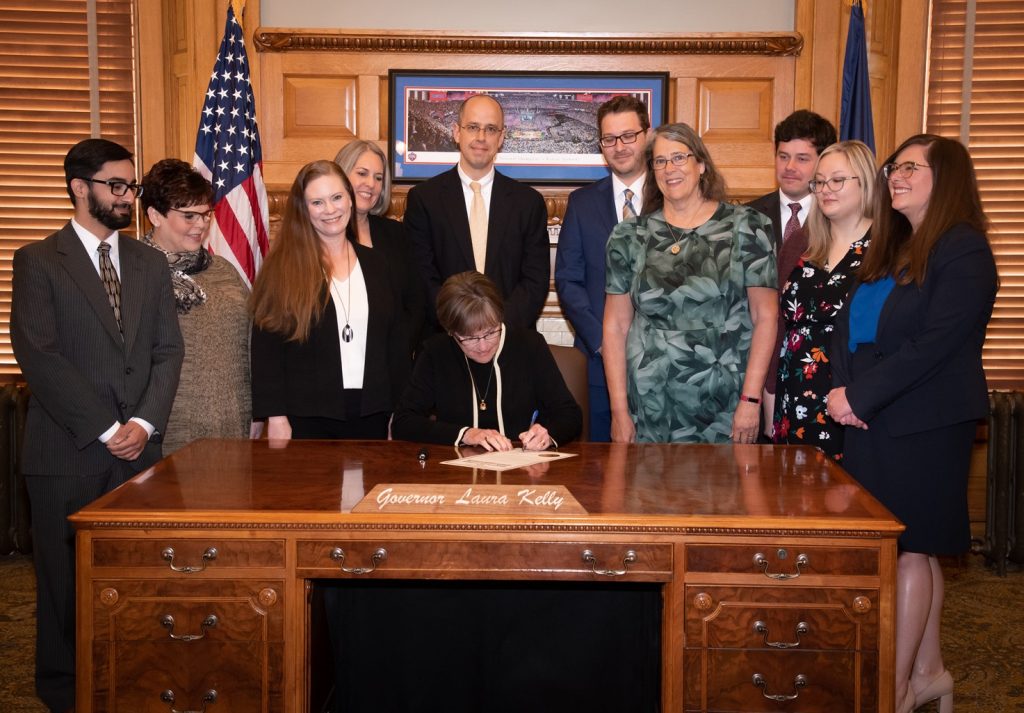 Harp, Kansas Legal Services staff, and Kansas Bar Association staff and board members gather around Laura Kelly's desk as she signs a proclamation for KLS's 45th anniversary.
